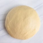 A round ball of smooth, raw brioche dough rests on a white, lightly marbled surface.