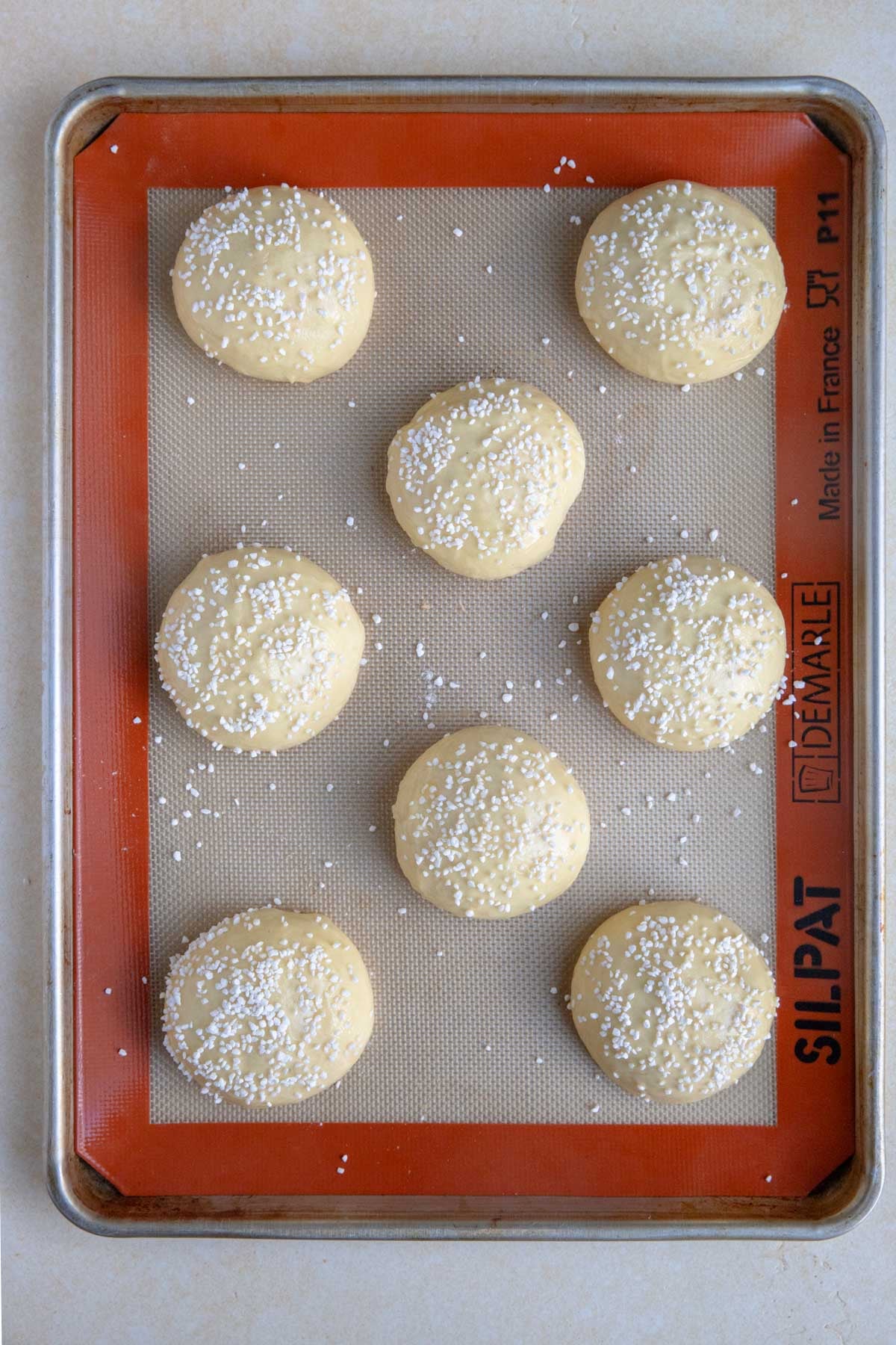 Eight pieces of dough rolled into balls arranged on a baking sheet lined with Silpat. Each piece of dough is brushed with egg wash and topped with Swedish pearl sugar