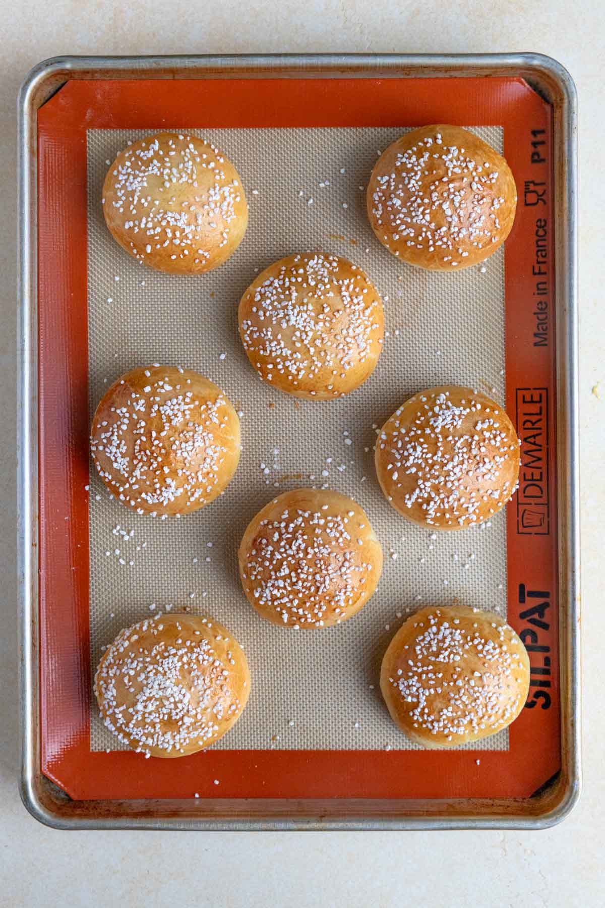 Eight baked brioche au sucre buns arranged on a baking sheet lined with Silpat. Each bun is topped with Swedish pearl sugar