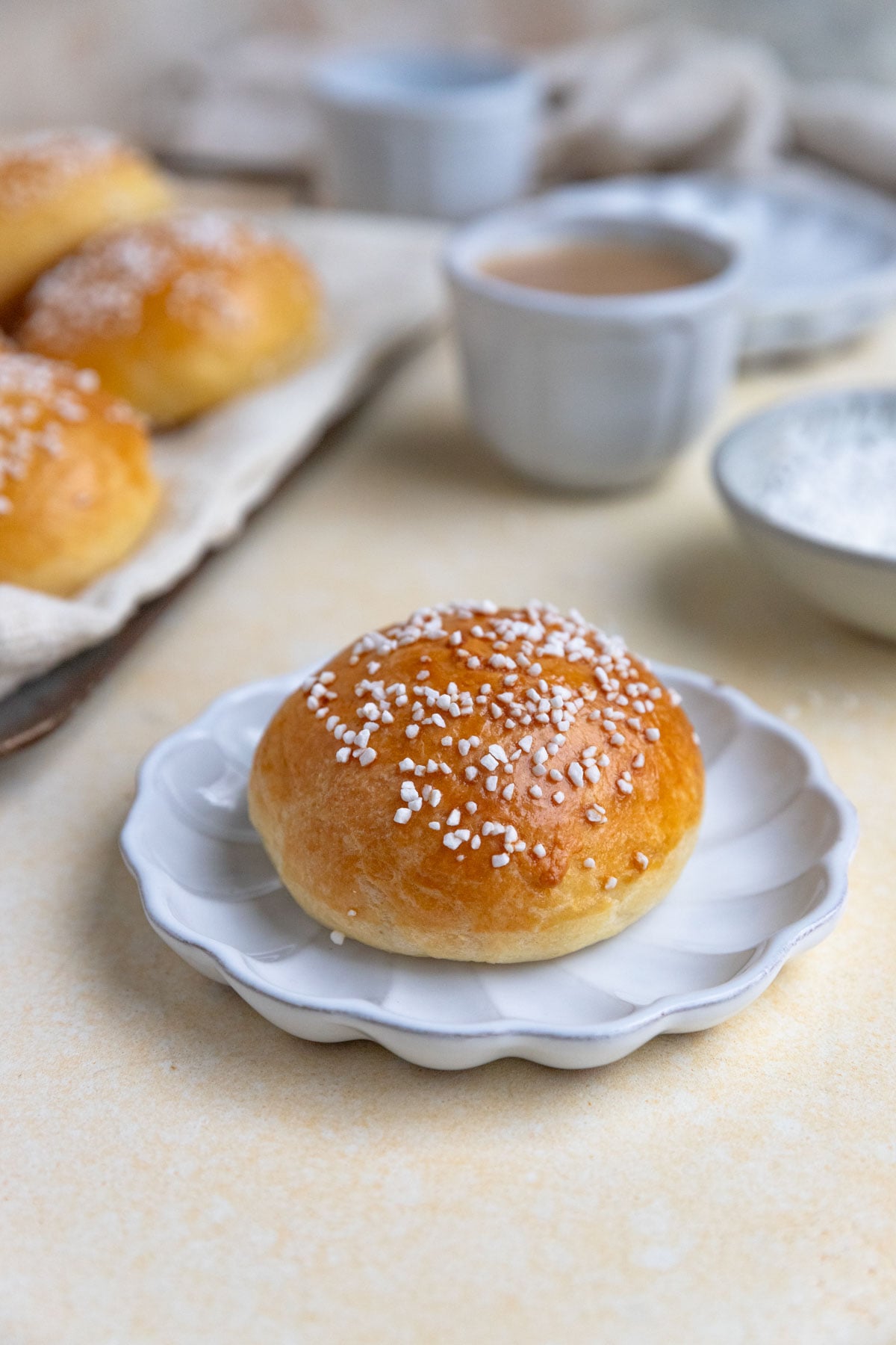 A brioche au sucre bun on a small white plate. There is a small cup of coffee in the background and a tray with a few brioche buns.