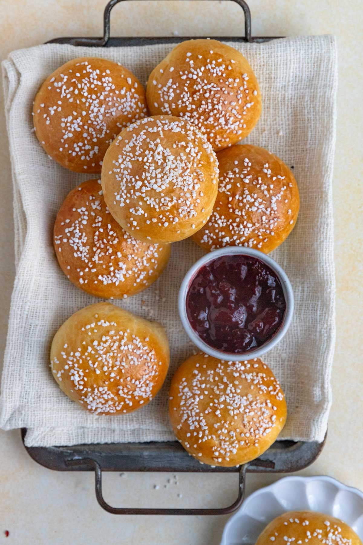 Seven brioche au sucre buns arranged on a platter lined with a kitchen towel. There is a small bowl of strawberry jam nestled among the buns.