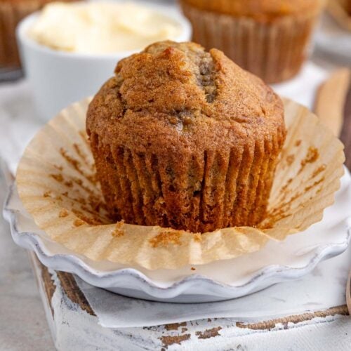 Banana muffin on a small plate with the muffin liner peeled down. A small bowl of butter is behind the muffin with a wooden knife next to the muffin