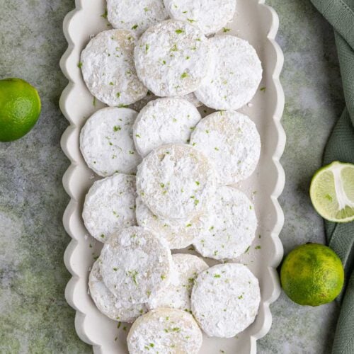 Several lime shortbread cookies with powdered sugar arranged on a long rectangular platter with fresh limes near the platter