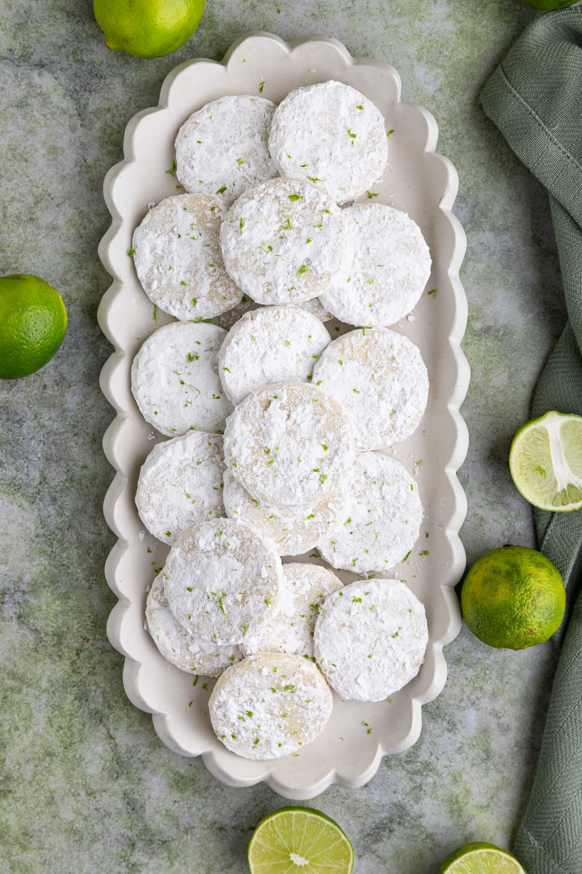 Several lime shortbread cookies with powdered sugar  arranged on a long rectangular platter with fresh limes near the platter
