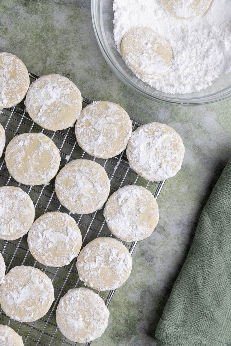 Lime shortbread cookies covered with powdered sugar on a wire cooling rack.  There is a small bowl with powdered sugar and a couple of cookies next to the wire rack.