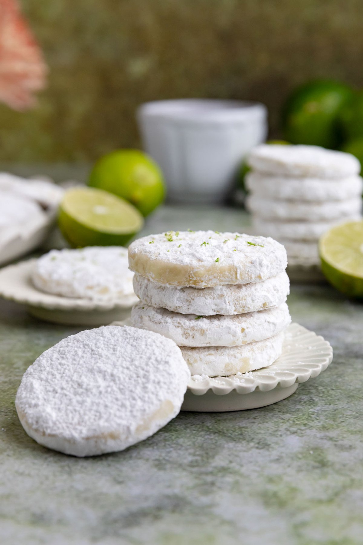 Two small stacks of lime shortbread cookies on two small plates with a teacup and small limes in the background.