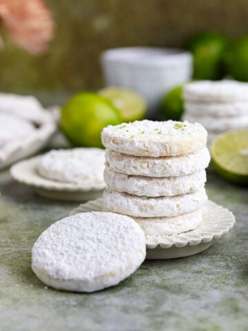 Two small stacks of lime shortbread cookies on two small plates with a teacup and small limes in the background.