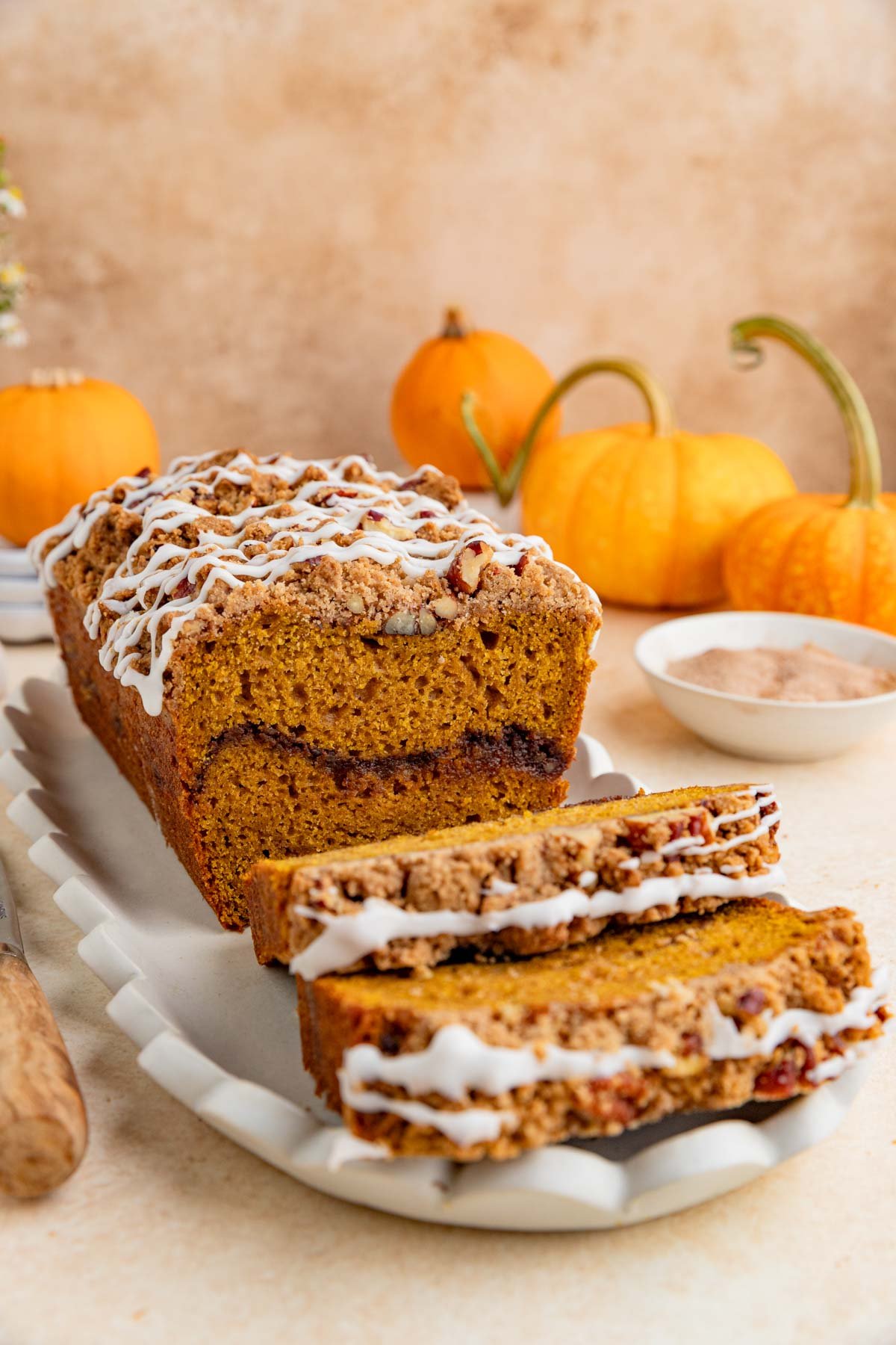 A loaf of pumpkin bread with a pecan streusel topping and white icing sits on a rectangular platter. Two slices are cut in front of the loaf. Small pumpkins and a bowl of cinnamon sugar are in the background.