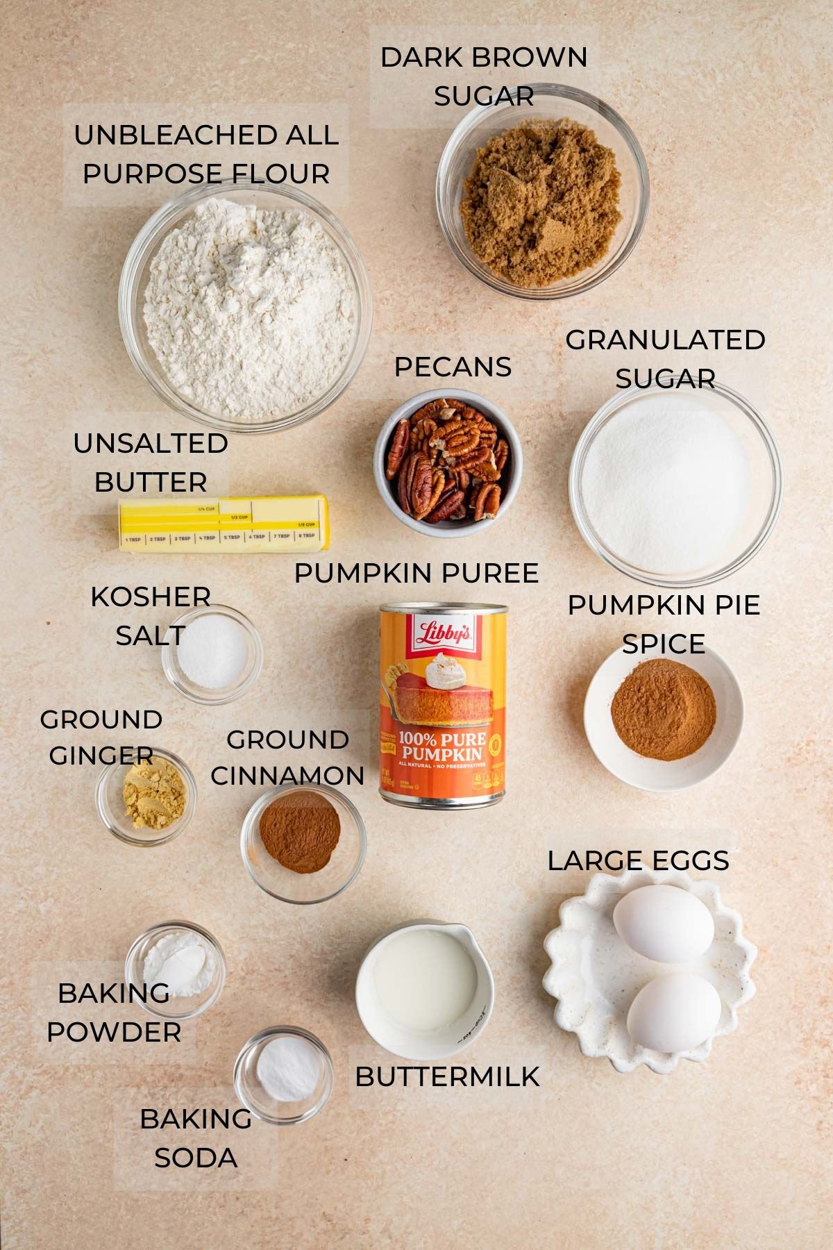 Recipe Ingredients arranged on a flat surface - dark brown sugar, pecans, granulated sugar, pumpkin pie spice, eggs, buttermilk, baking soda, baking powder, ground cinnamon, ground ginger, kosher salt, a can of pumpkin puree, unsalted butter, all-purpose flour