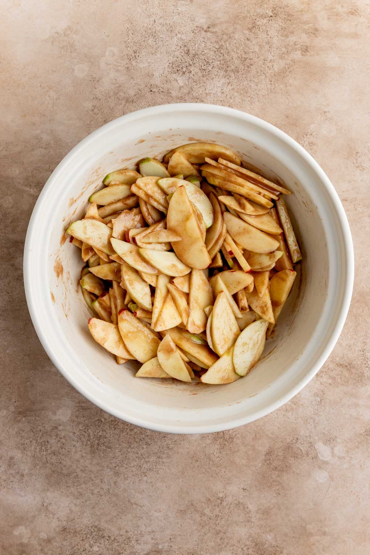 Apples mixed with sugar, flour, cinnamon, nutmeg, cardamom, and salt in a large off-white ceramic bowl