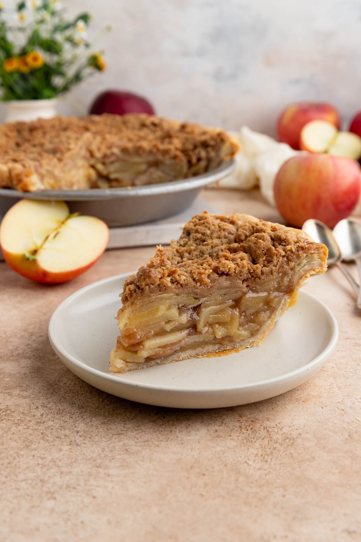 A slice of apple crumble pie on a small plate. The rest of the pie and some fresh apples are in the background