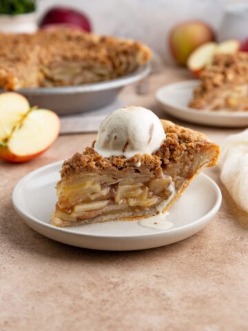A slice of apple crumble pie on a small plate with a scoop of ice cream on top. The rest of the pie and some fresh apples are in the background