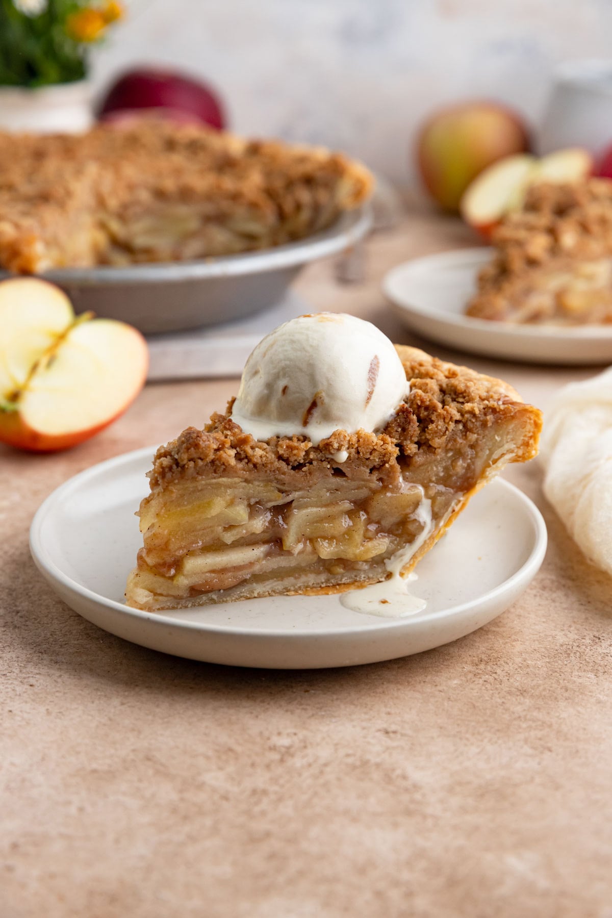 A slice of apple crumble pie on a small plate with a scoop of ice cream on top.  The rest of the pie and some fresh apples are in the background