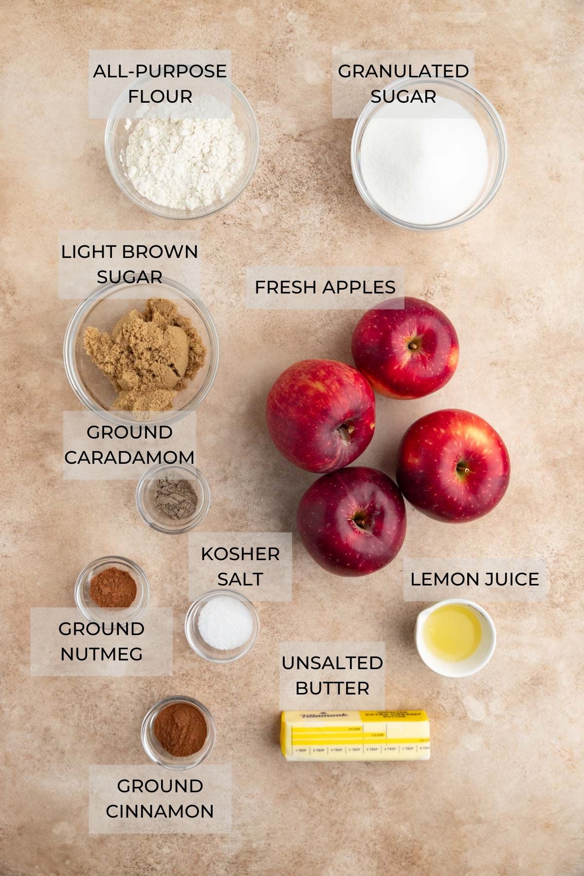 Apple Crumble Pie ingredients arranged on a light brown board. Clockwise: granulated sugar, fresh apples, lemon juice, butter, ground cinnamon, ground nutmeg, kosher salt, ground cardamom, light brown sugar, AP flour