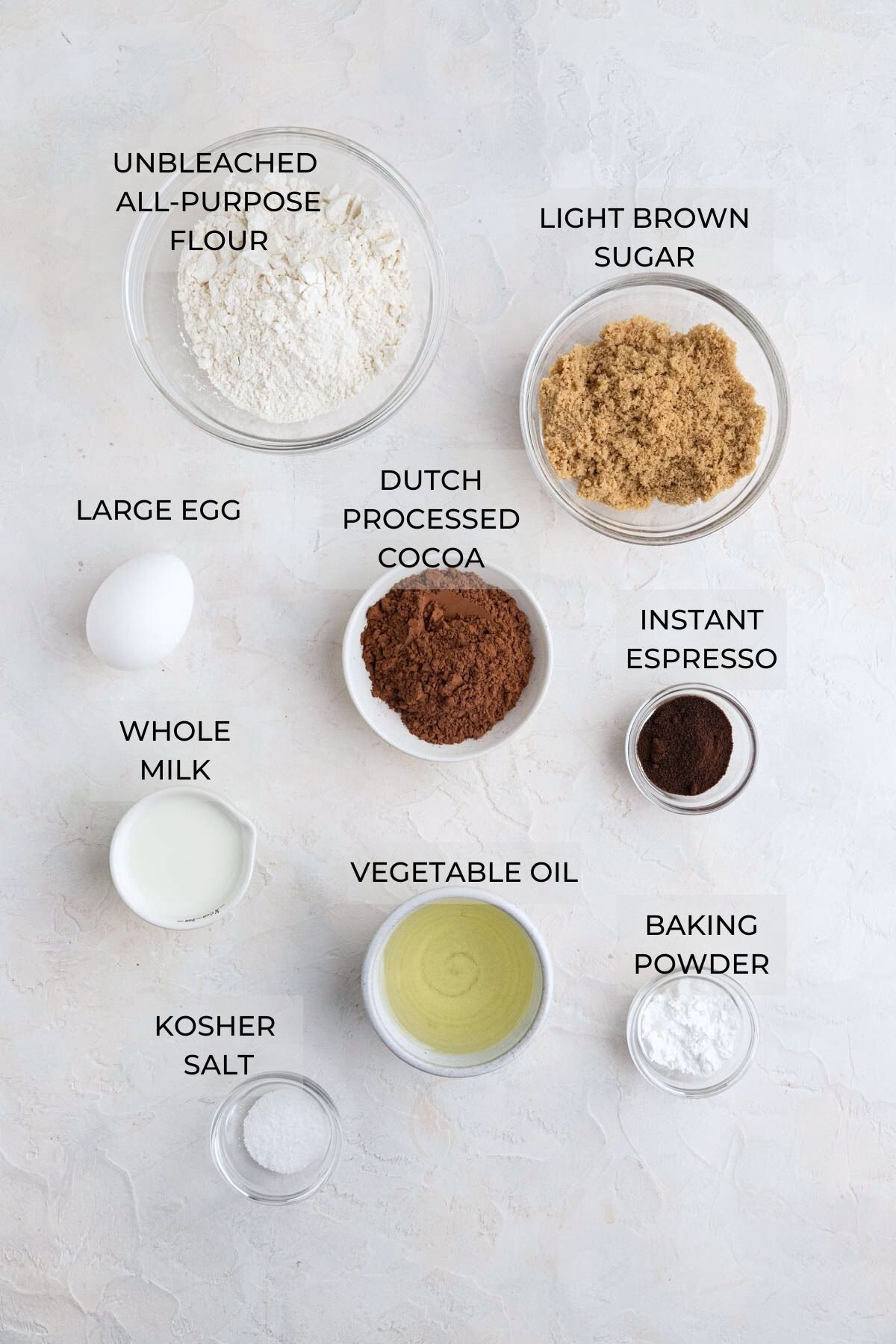Chocolate doughnut ingredients arranged on a white board.  Clockwise: light brown sugar, Dutch processed cocoa, espresso powder, baking powder, vegetable oil, kosher salt, whole milk, an egg, and unbleached all-purpose flour