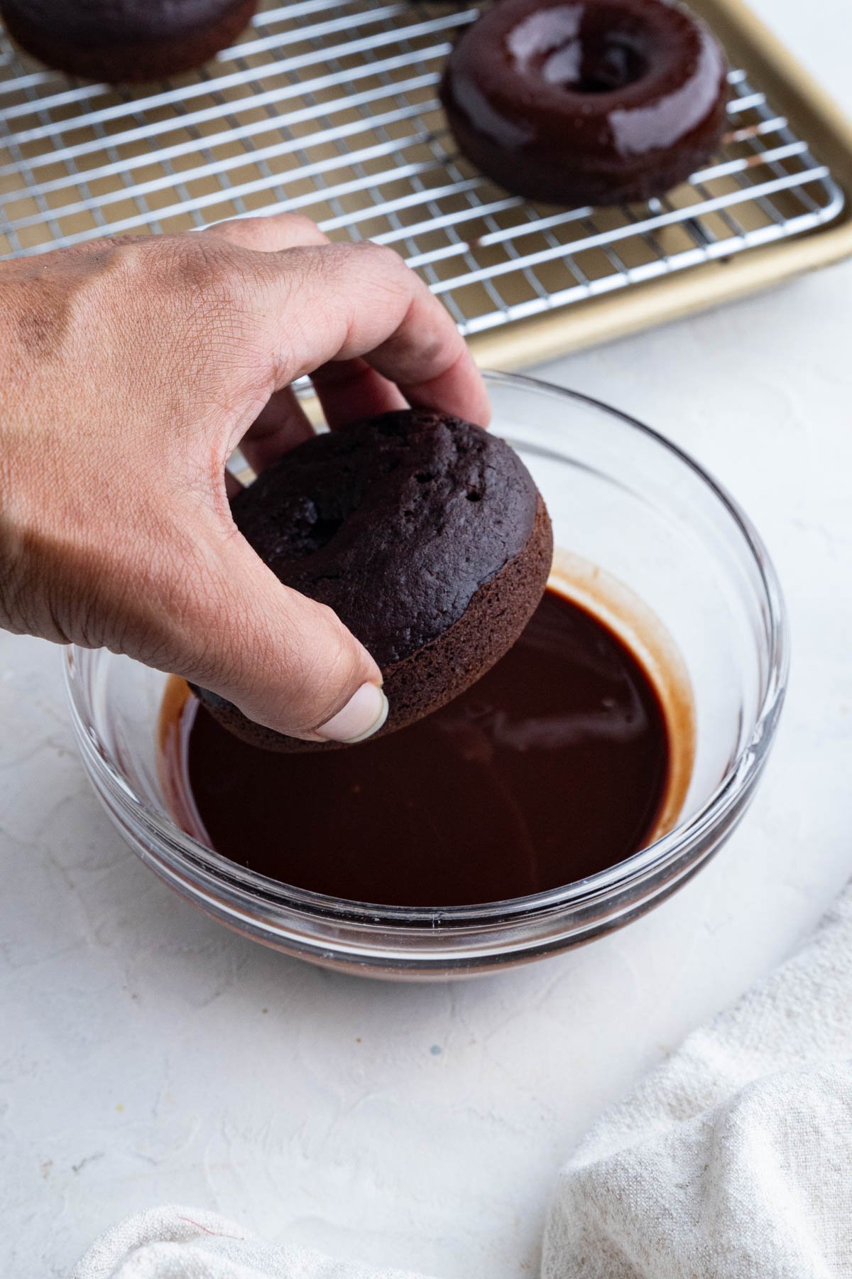 A hand dips a chocolate donut into a bowl of chocolate glaze. In the background, glazed donuts cool on a wire rack set on a baking tray. A white towel is partially visible in the lower right corner.