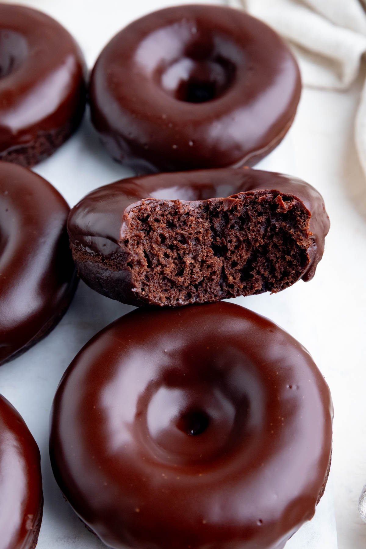 A close-up of several chocolate glazed donuts, with one donut resting on top of another and a bite taken out, showing the soft, chocolate interior. The surface of the doughnuts is shiny and smooth.