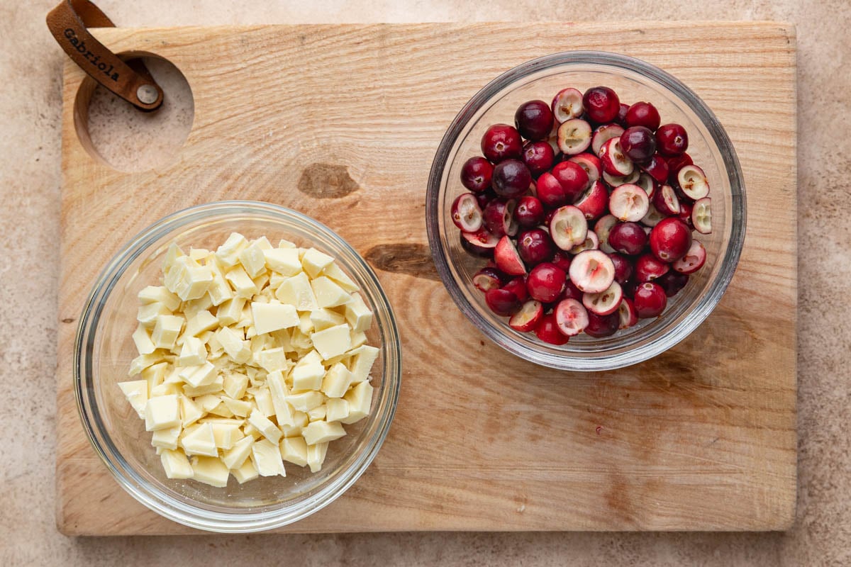 Two small bowls on a wooden board. Once is filled with chopped white chocolate and the other is filled with freshed chopped cranberries