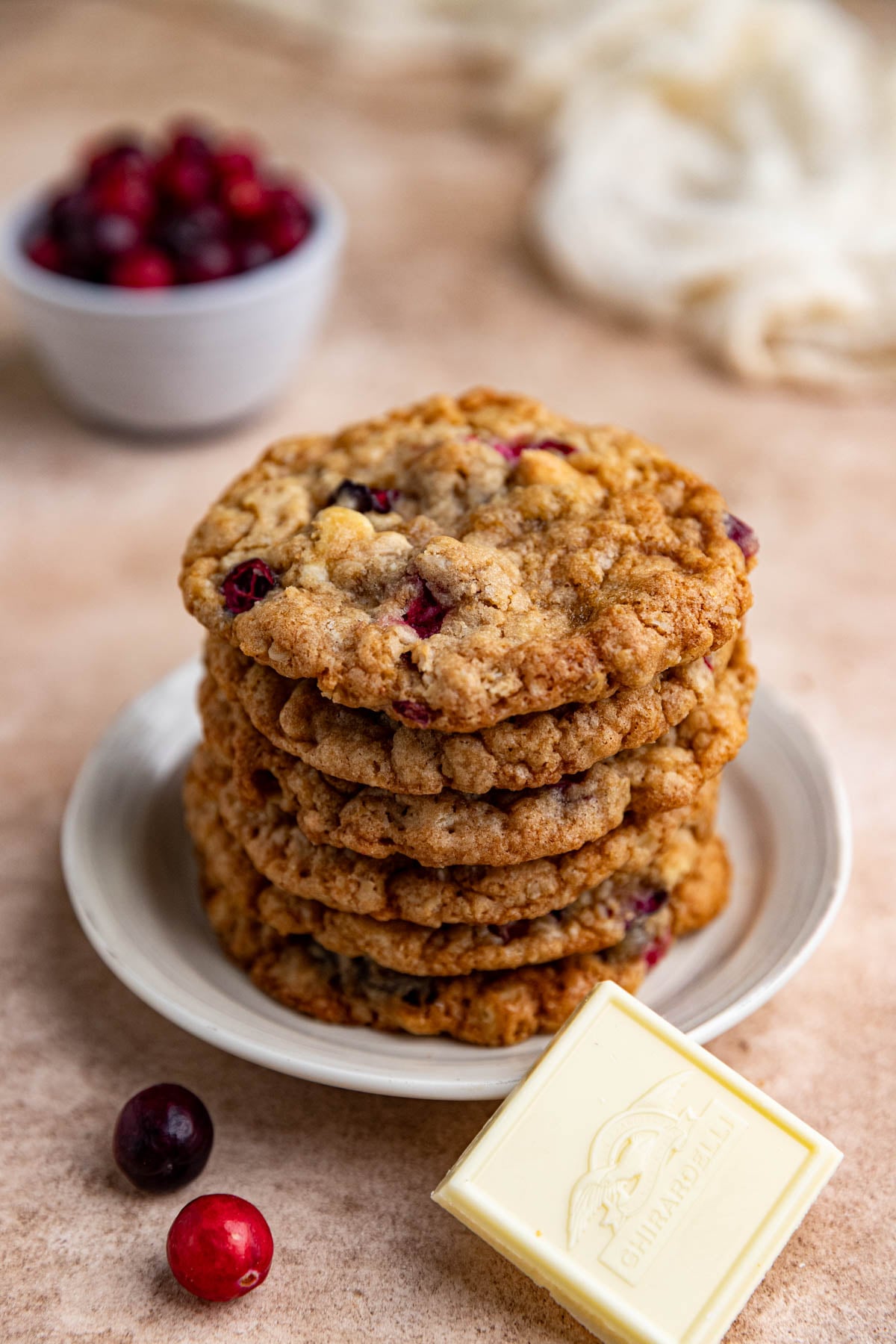 Six cranberry white chocolate oatmeal cookies stacked on a small white plate with a small bowl of cranberries in the background