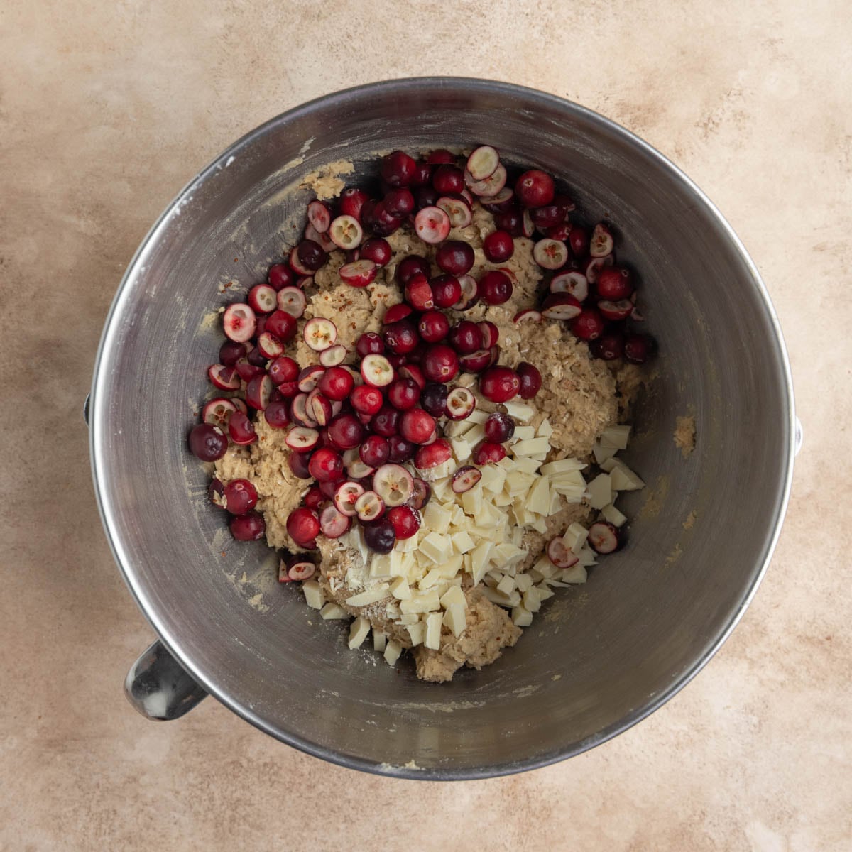 The chopped cranberries and chopped white chocolate on top of the cookie dough in a stainless steel bowl
