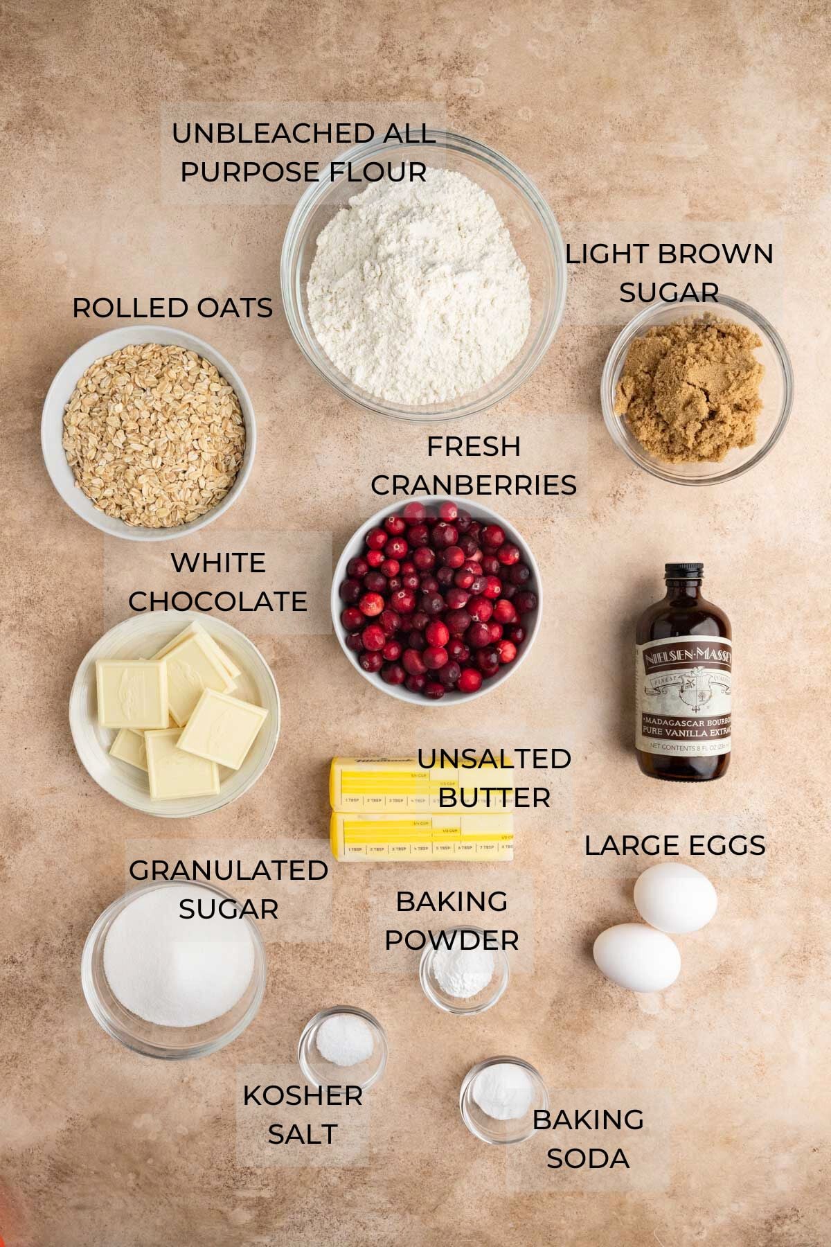 Cranberry White Chocolate Oatmeal Cookie ingredients arranged on a brown board in glass prep bowls - all purpose flour, light brown sugar, vanilla extract, large eggs, unsalted butter, baking powder, baking soda, kosher salt, granulated sugar, white chocolate, rolled oats