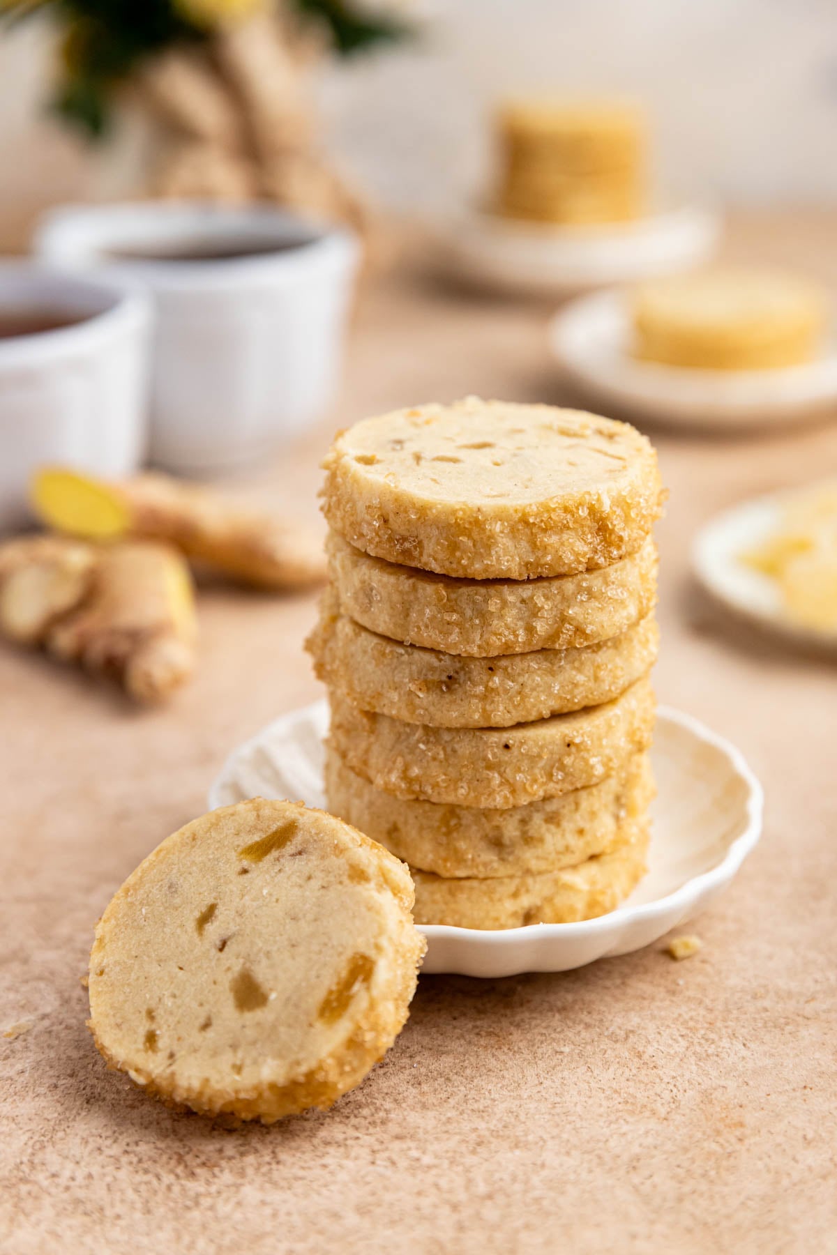 A stack of 6 ginger shortbread cookies on a small white plate, with one cookie leaning against the plate. There are two cups of tea in the background.