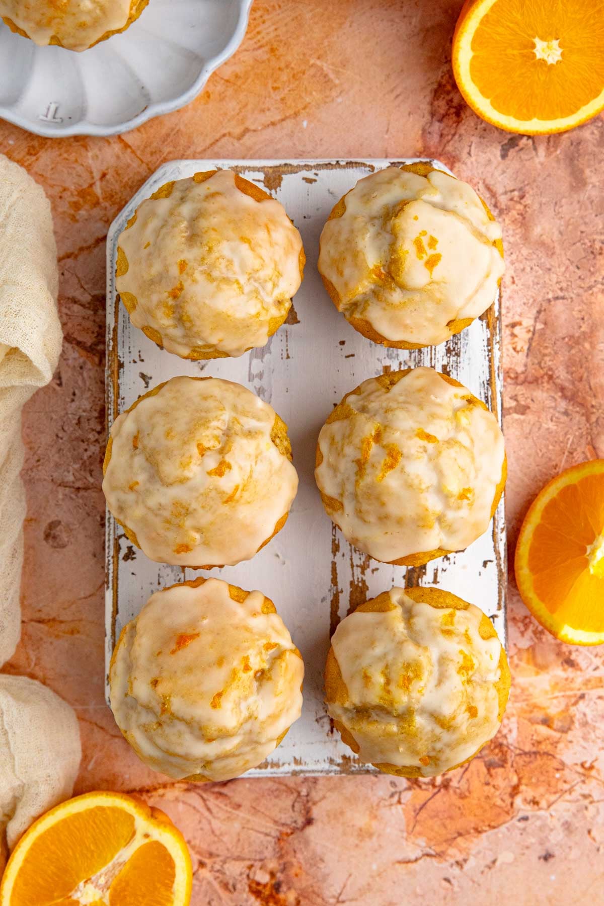 A top down view of six orange muffins with orange glaze arranged on a small wooden board. There are fresh oranges near the board