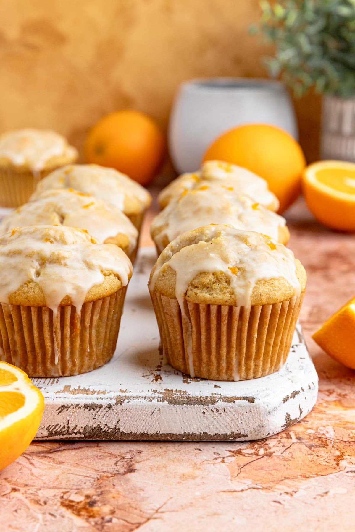 Six orange muffins with orange glaze arranged on a small wooden board.  There are fresh oranges in the background.