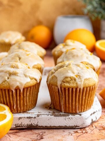 Six orange muffins with orange glaze arranged on a small wooden board. There are fresh oranges in the background.