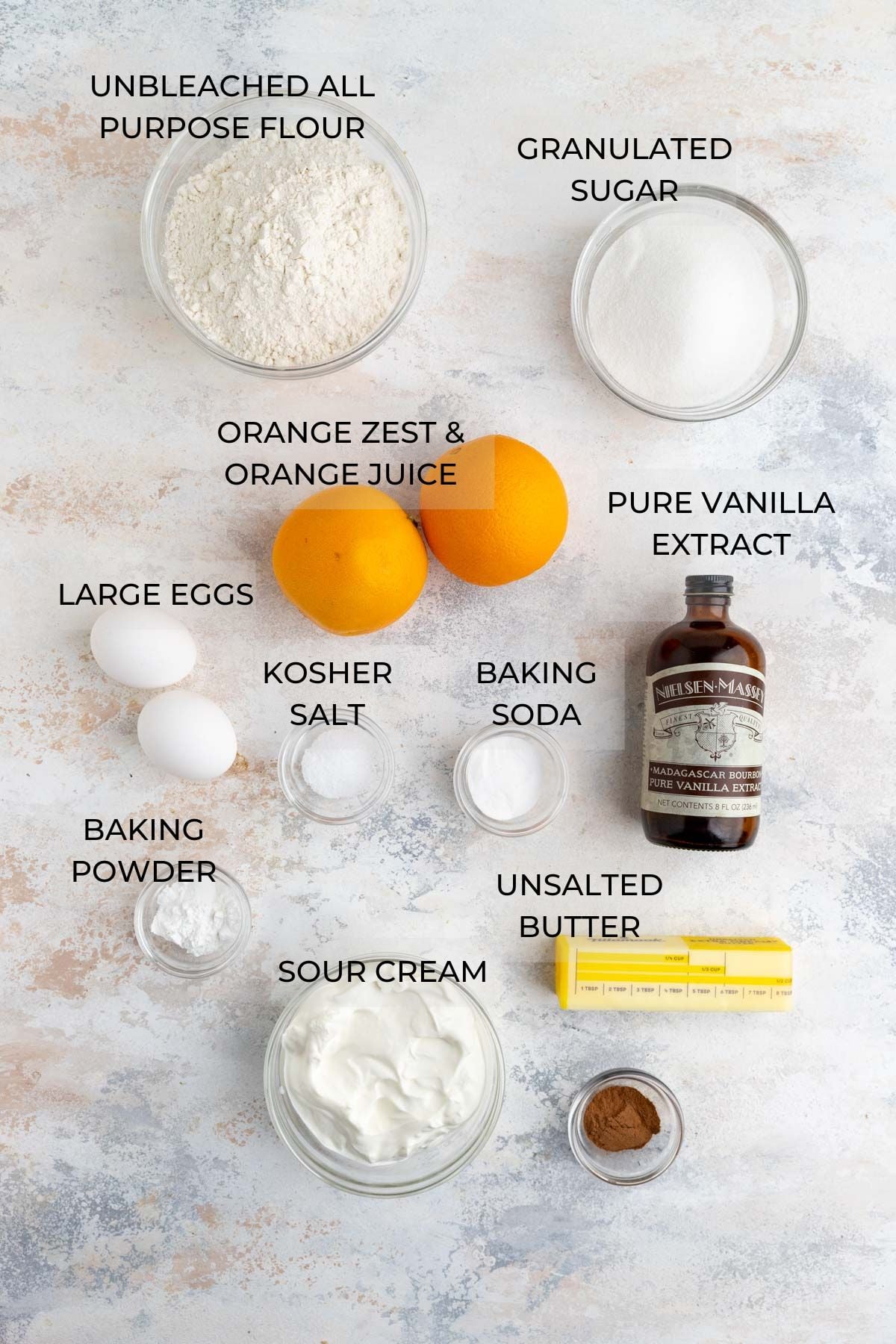Orange muffin ingredients arranged on a board - unbleached all-purpose flour, granulated sugar, vanilla extract, unsalted butter, ground cinnamon, sour cream, baking soda, kosher salt, baking powder, eggs, and oranges