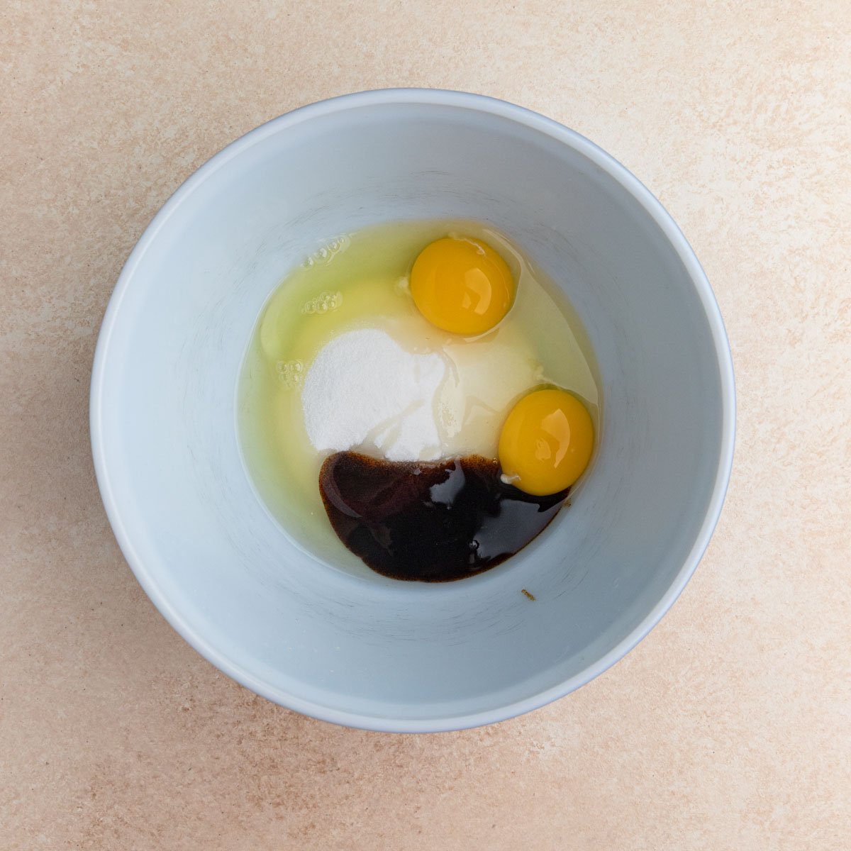 Two eggs, sugar, and vanilla bean paste in a white ceramic bowl.