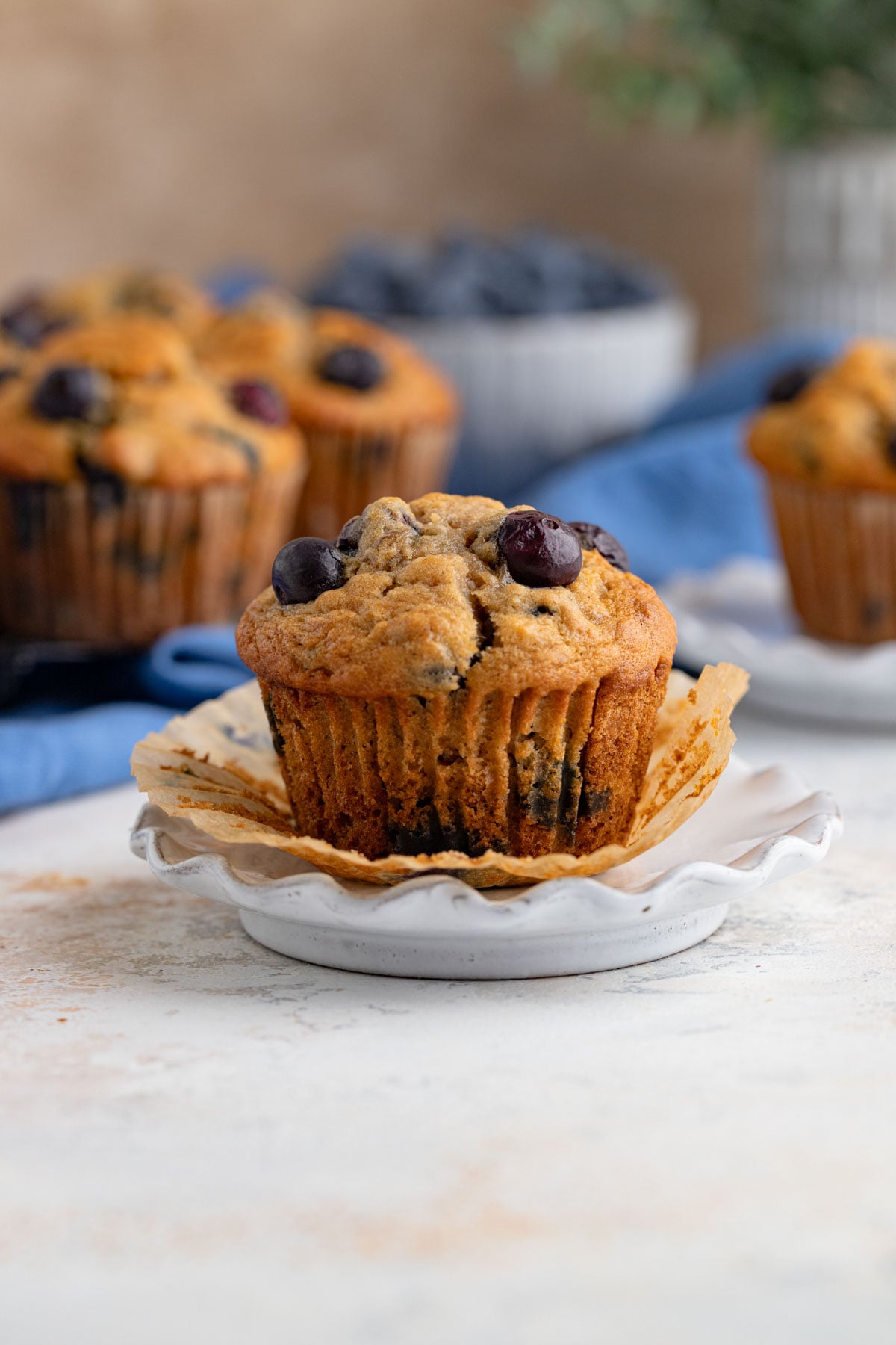 A single blueberry banana muffin on a small plate with the muffin wrapper removed.  More muffins are in the background with a small bowl of blueberries