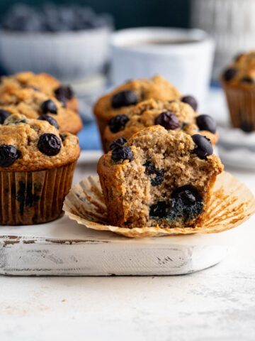 Six blueberry banana muffins on a white board with a bowl of blueberries in the background. A muffin in the front has a bite taken out of it.