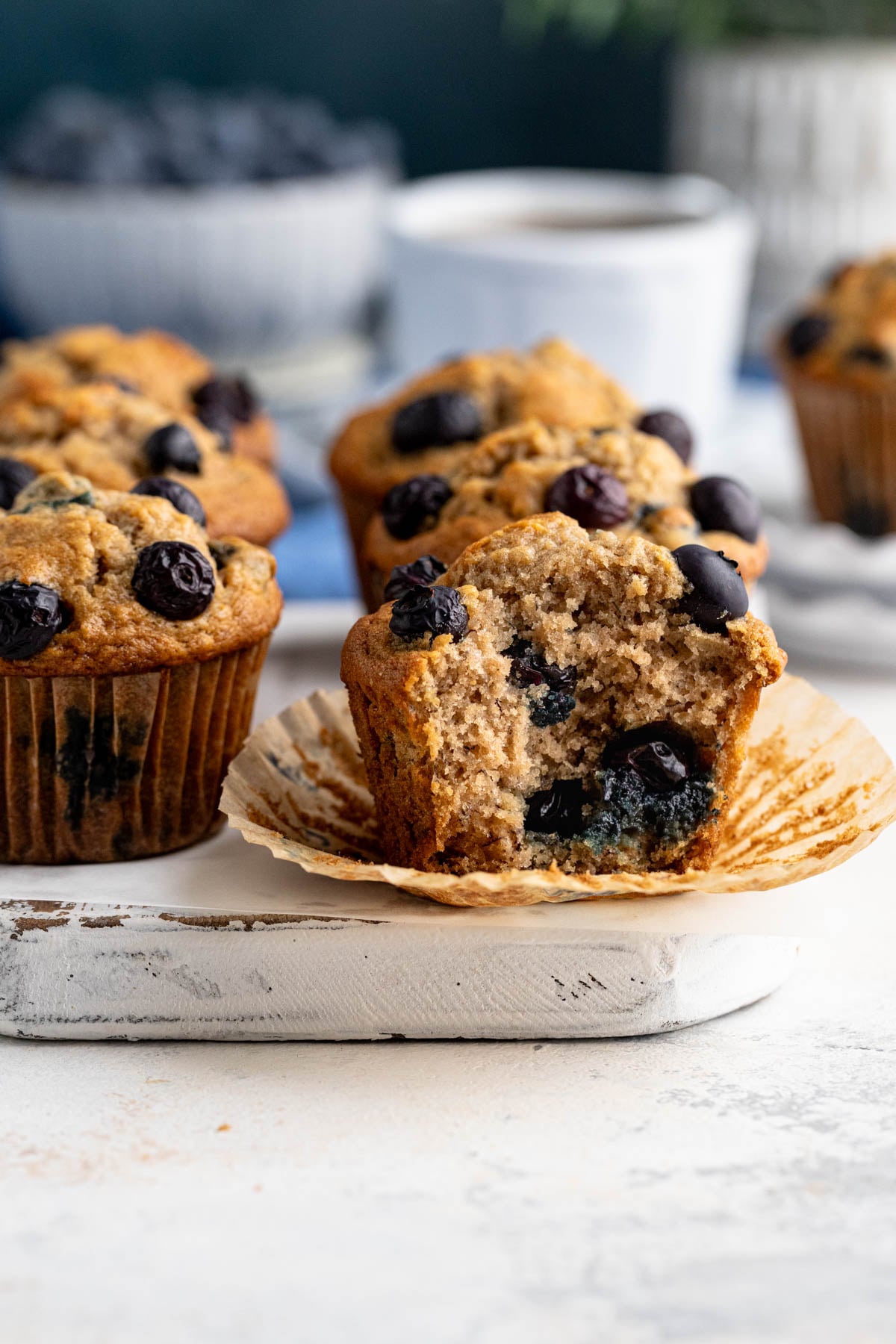 Six blueberry banana muffins on a white board with a bowl of blueberries in the background. A muffin in the front has a bite taken out of it.