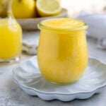 Lemon curd in an 8-ounce jar on a small white plate. A small container of lemon juice and fresh lemons are in the background