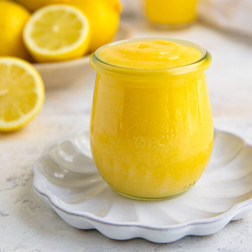 Lemon curd in an 8-ounce jar on a small white plate. Fresh lemons are in the background.