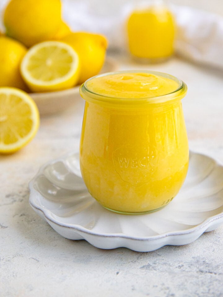 Lemon curd in an 8-ounce jar on a small white plate. Fresh lemons are in the background.