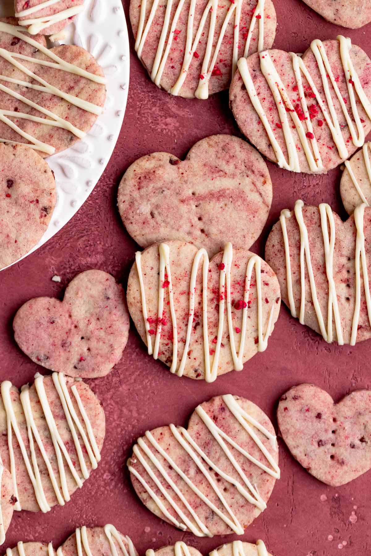 Raspberry shortbread cookies on a red surface with some of the cookies on a white plate.  The cookies are covered with a white chocolate glaze