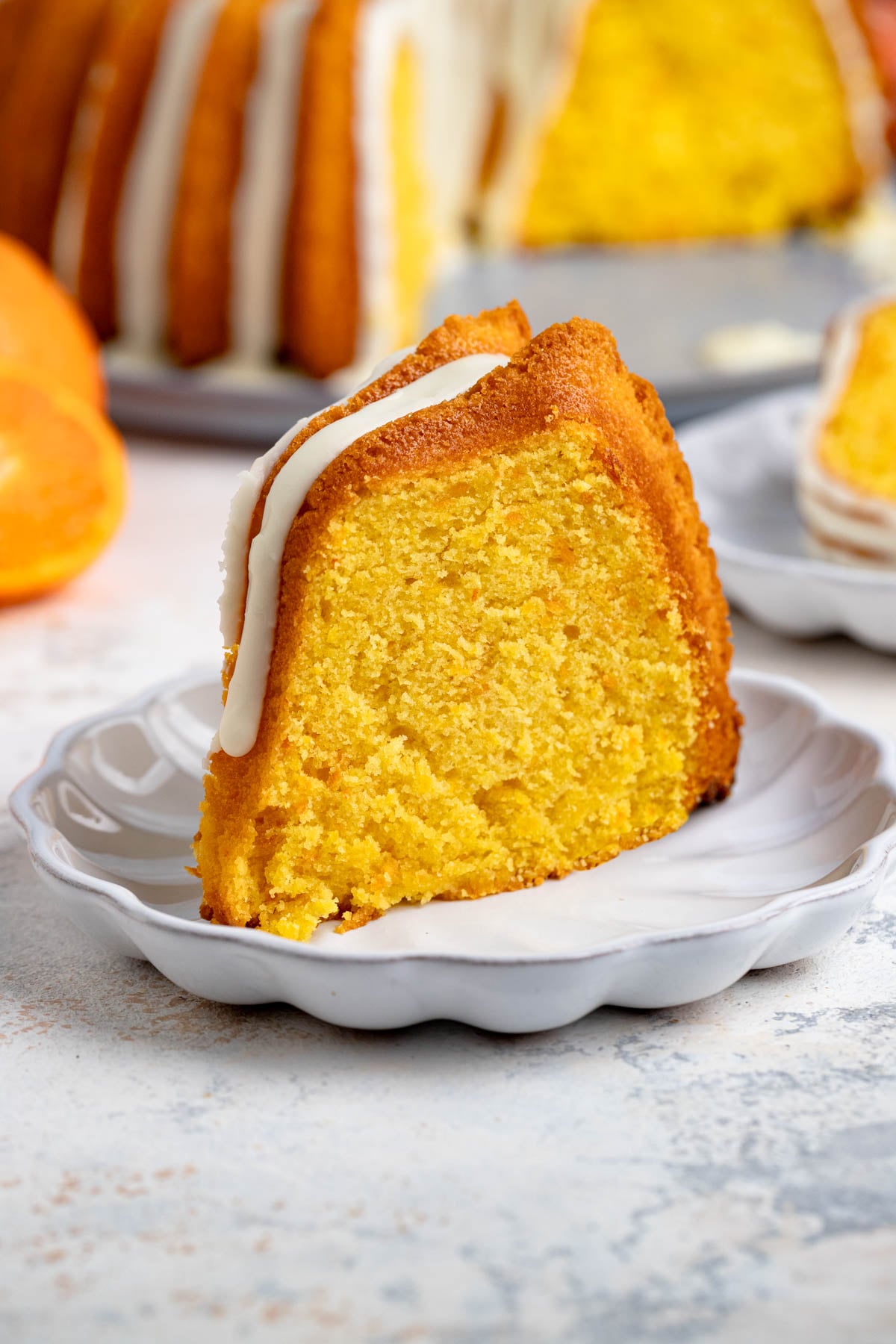 A single slice of Clementine cake on a small white dessert plate, with the rest of the cake in the background.