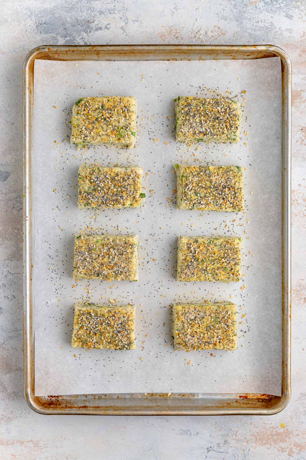 Eight unbaked scones on a parchment lined baking sheet. The top of the scones are covered with homemade everything bagel seasoning.