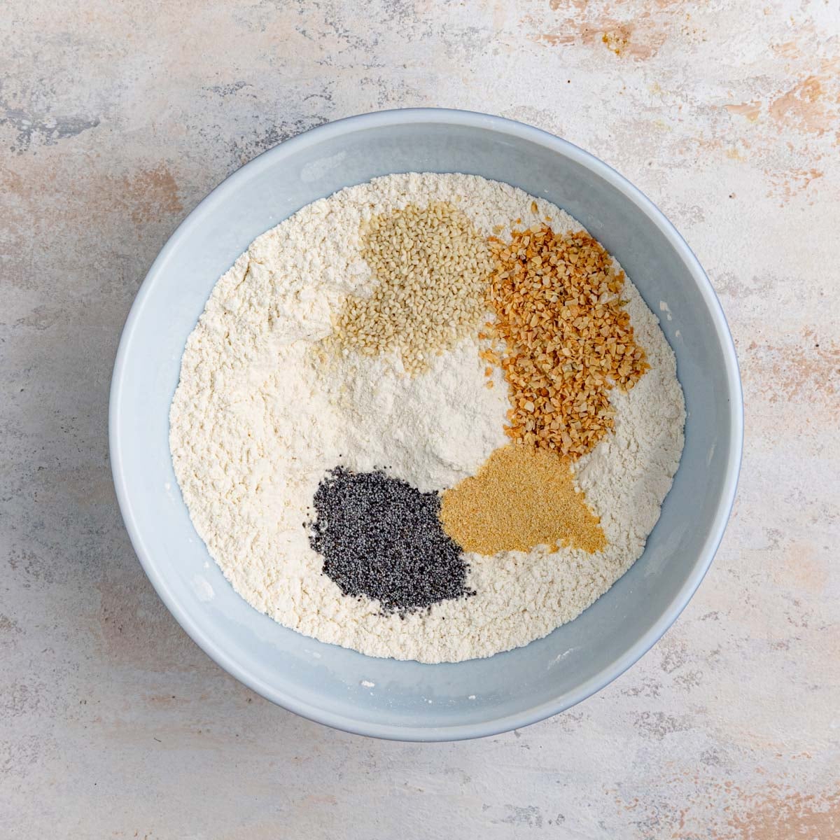 Flour mixture, poppy seeds, granulated garlic, dried onions, white sesame seeds in a white ceramic bowl