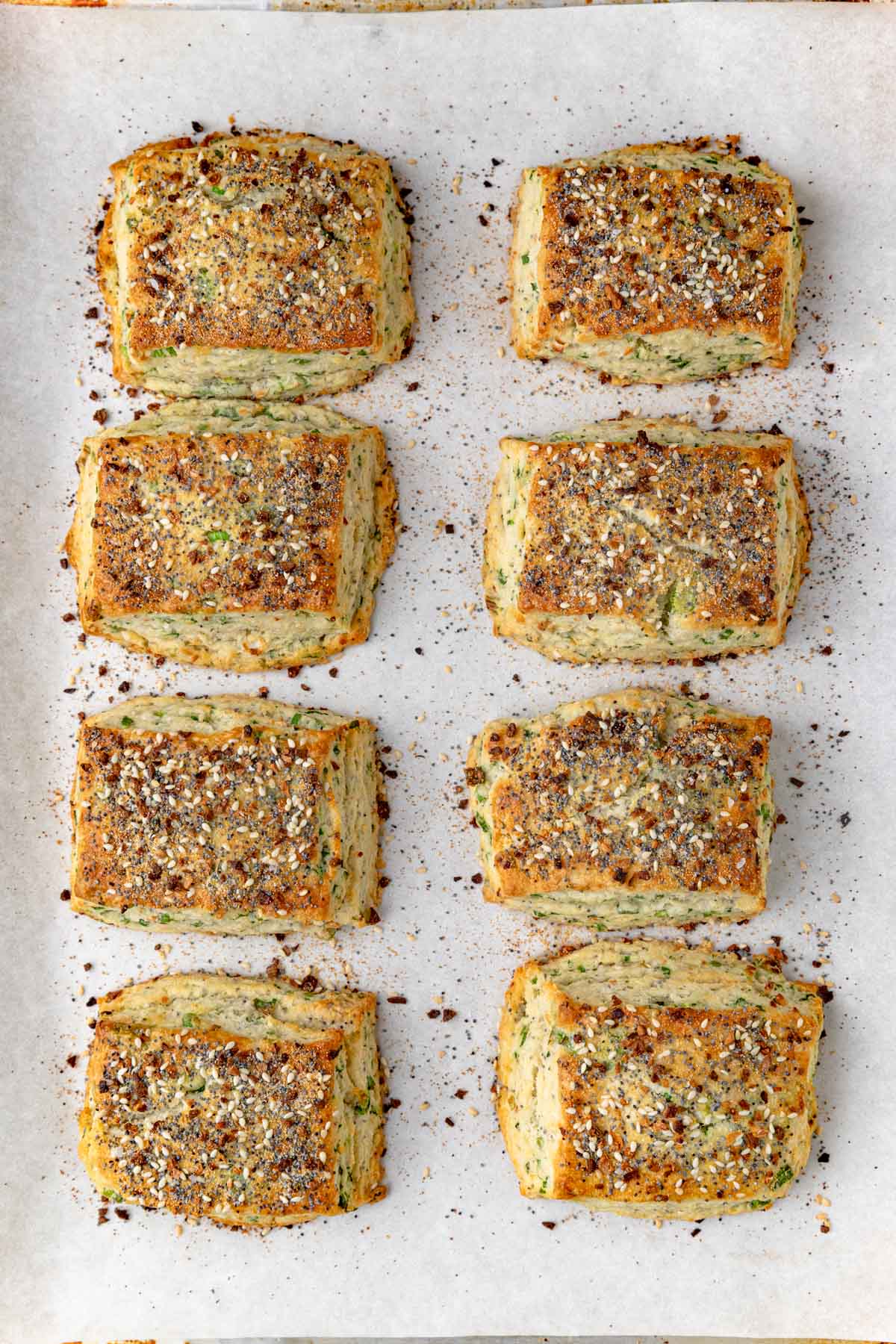 Eight rectangular, golden-brown scones topped with everything bagel seasoning are arranged in two rows of four on a sheet of parchment paper. The scones are flaky