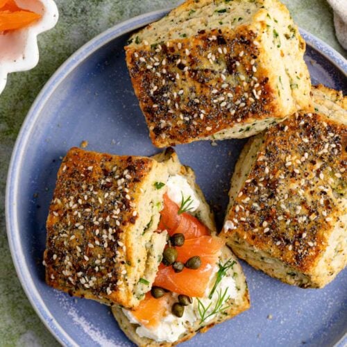 Three everything bagel scones on a blue plate. One scone is split in half and has cream cheese, smoked salmon, and capers spread on the bottom half.