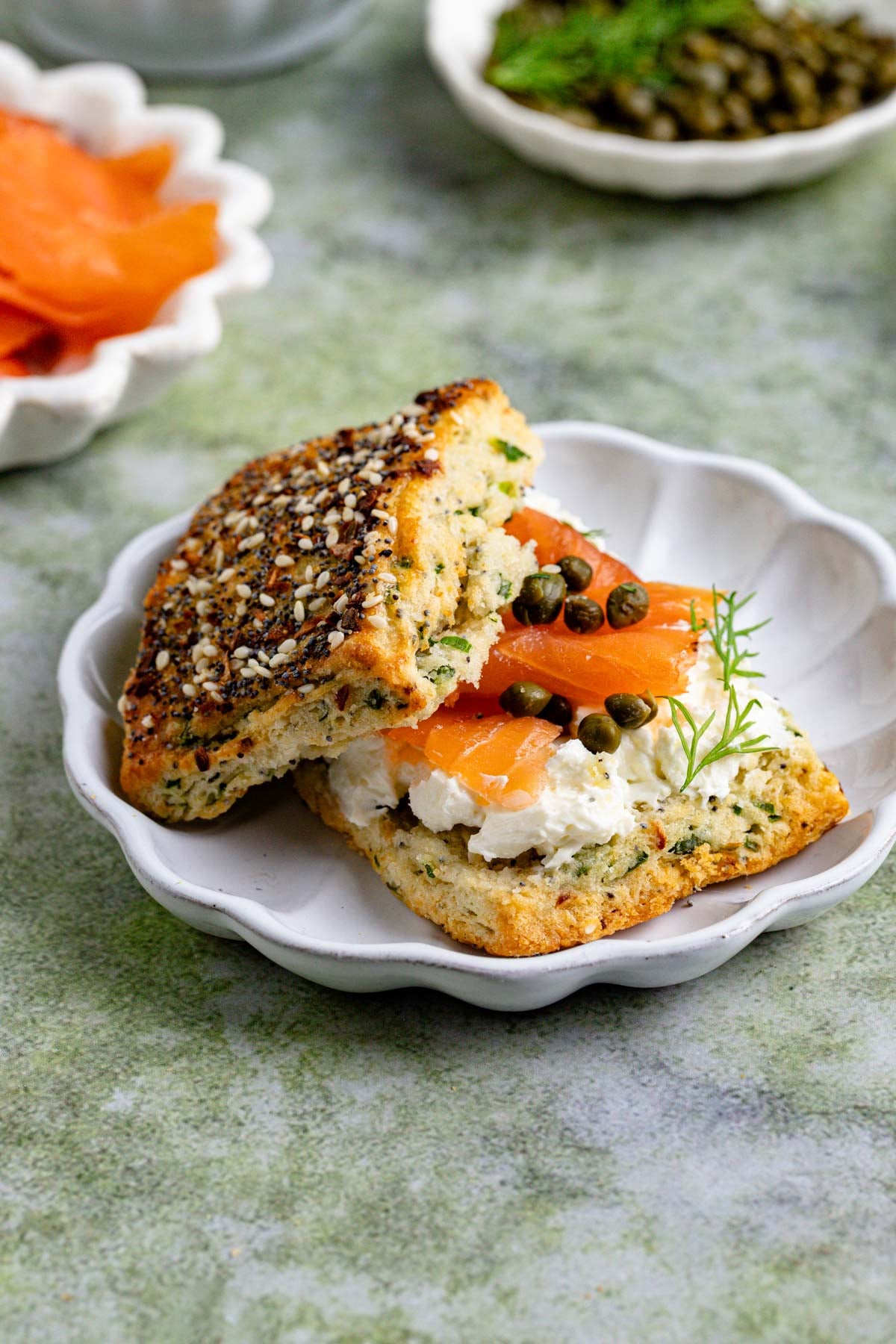 An everything bagel scone split in half with cream cheese, smoked salmon, capers, and dill on a white plate. In the background, there are small bowls with sliced smoked salmon, capers, and dill.