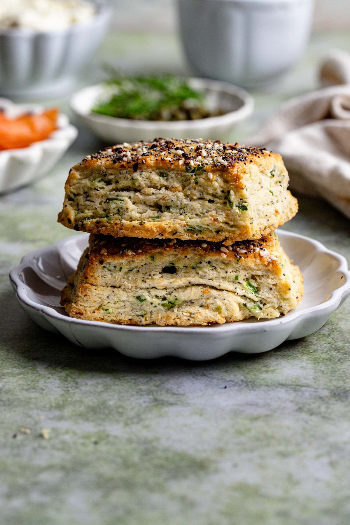 Two everything bagel scones stacked on a small white plate with whipped cream cheese, capers, smoked salmon in the background