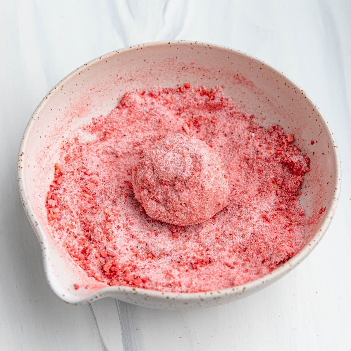 Strawberry sugar mixture in a creamed color bowl with one ball of dough in the middle of the mixture.