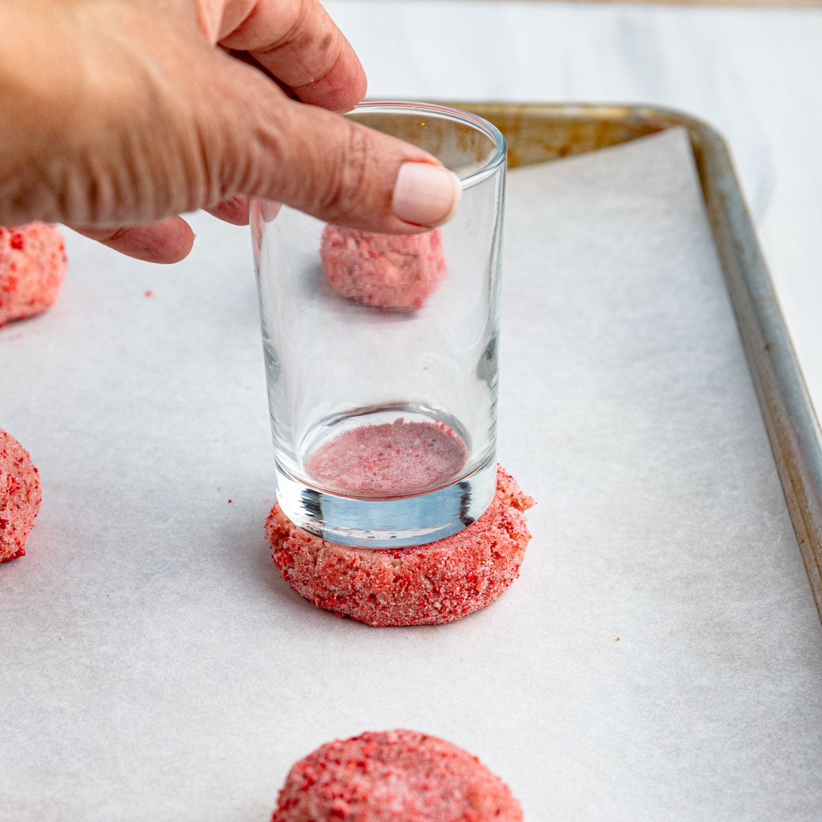 A hand pressing a round pink cookie dough ball flat with the bottom of a clear glass on a parchment-lined baking sheet. Other unflattened dough balls are visible in the background.