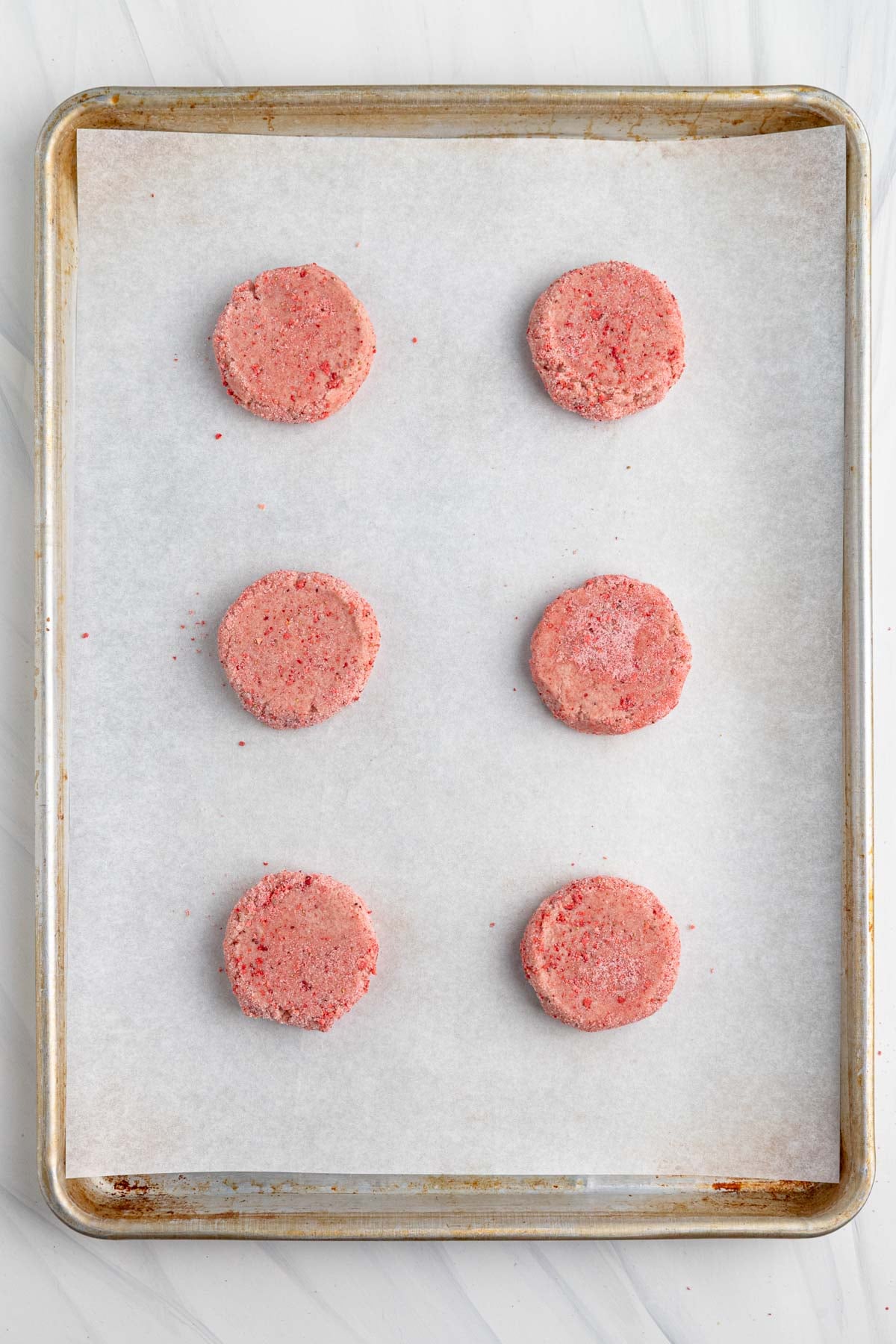 A baking sheet lined with parchment paper holds six evenly spaced, round, unbaked strawberry sugar cookies in columns of three.