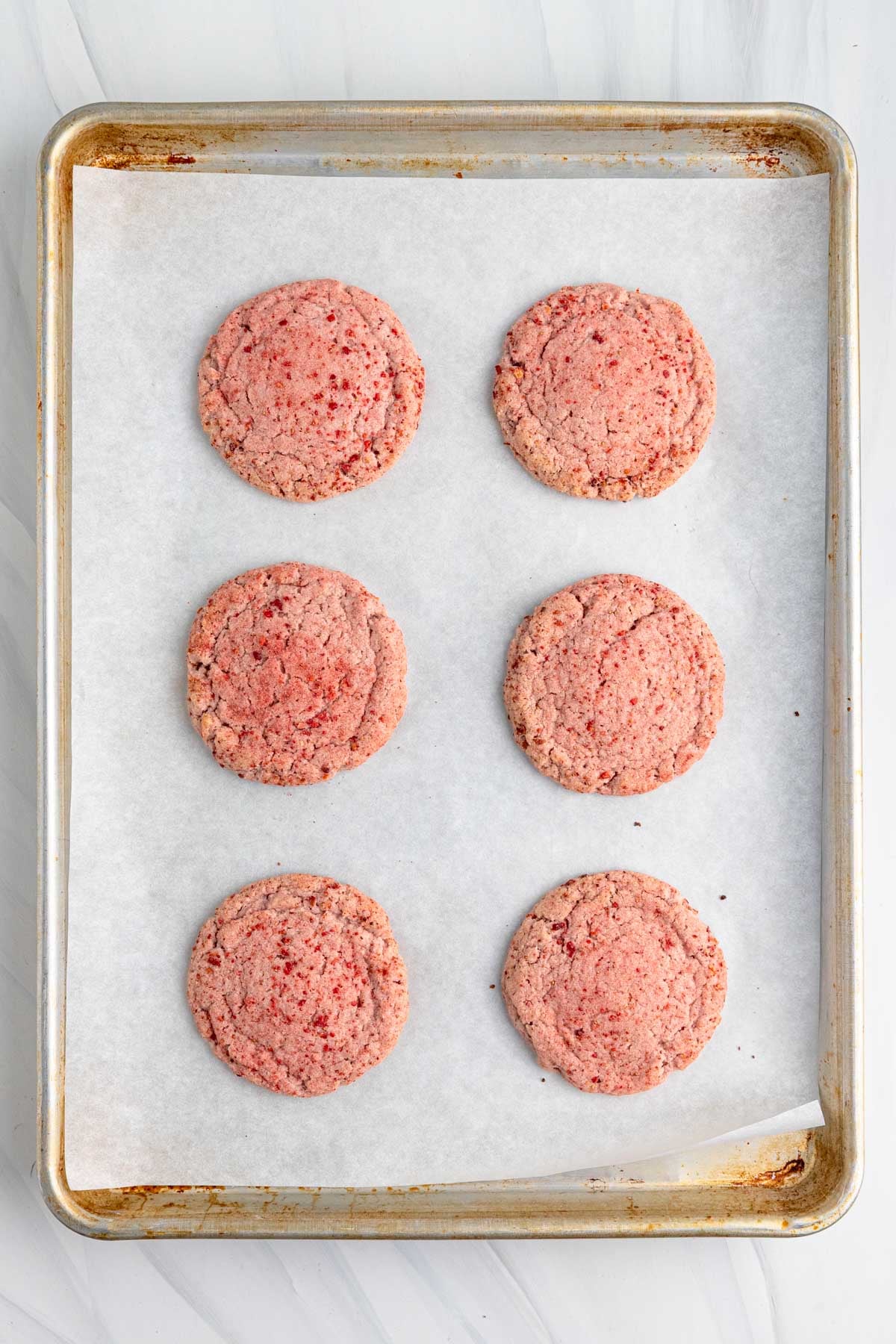 A baking sheet lined with parchment paper holds six evenly spaced, round, baked strawberry sugar cookies