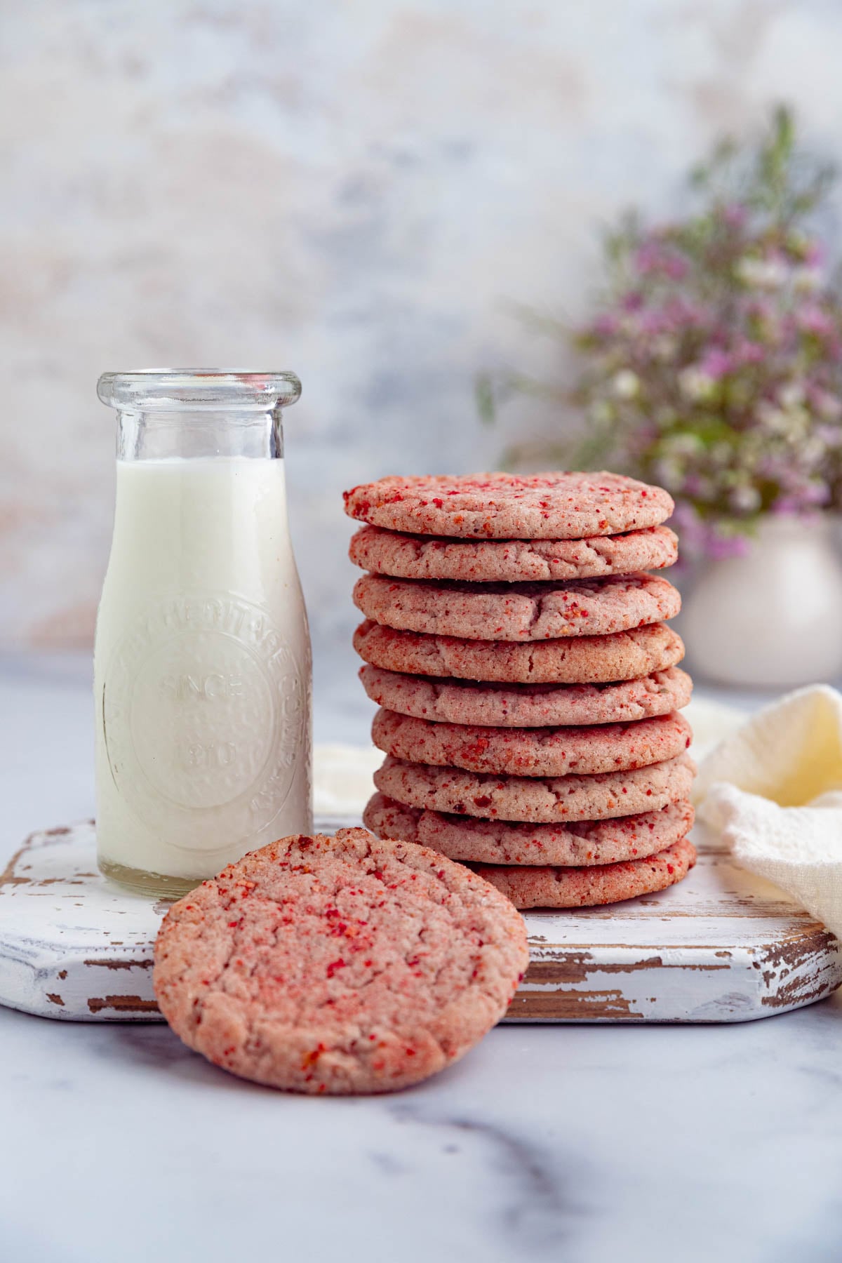 A stack of pink strawberry sugar cookies sits on a worn white wooden board next to a glass bottle of milk. One cookie is leaning against the stack. A bouquet of flowers is in the background.